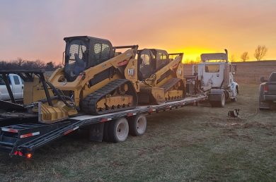 skid loaders on a truck and trailer with service vehicles parked near by.